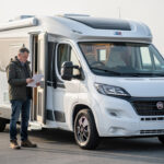 Camping-car profilé blanc stationné sur une aire de départ, avec un homme consultant des documents administratifs vierges près de la portière.
