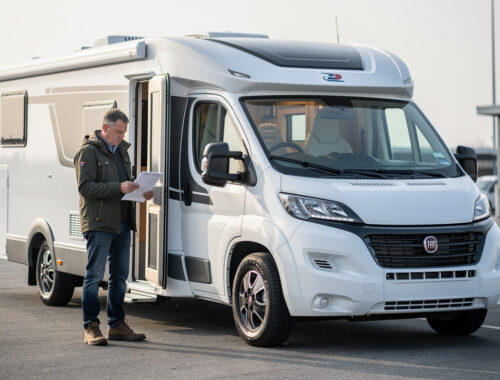 Camping-car profilé blanc stationné sur une aire de départ, avec un homme consultant des documents administratifs vierges près de la portière.