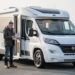 Camping-car profilé blanc stationné sur une aire de départ, avec un homme consultant des documents administratifs vierges près de la portière.
