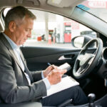 Un homme consulte son budget automobile dans l’habitacle d’une voiture moderne, avec une station-service visible en arrière-plan sous une lumière naturelle.