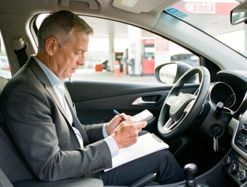 Un homme consulte son budget automobile dans l’habitacle d’une voiture moderne, avec une station-service visible en arrière-plan sous une lumière naturelle.