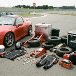 Équipement complet de sport automobile amateur soigneusement disposé dans le paddock d’un circuit, avec voiture de course, casque, combinaison, outils, pneus et caisses métalliques, sans pilote.