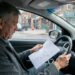 Un homme d’âge mûr consulte des feuilles vierges au volant d’une voiture stationnée dans une rue urbaine.