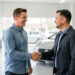 Un conseiller automobile accueille chaleureusement un client dans un garage moderne et lumineux, avec une voiture visible à l’arrière-plan.