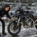 Une journaliste moto examine un trail chinois moderne dans une usine d’assemblage propre, avec des chaînes de production et des postes de contrôle qualité en arrière-plan.